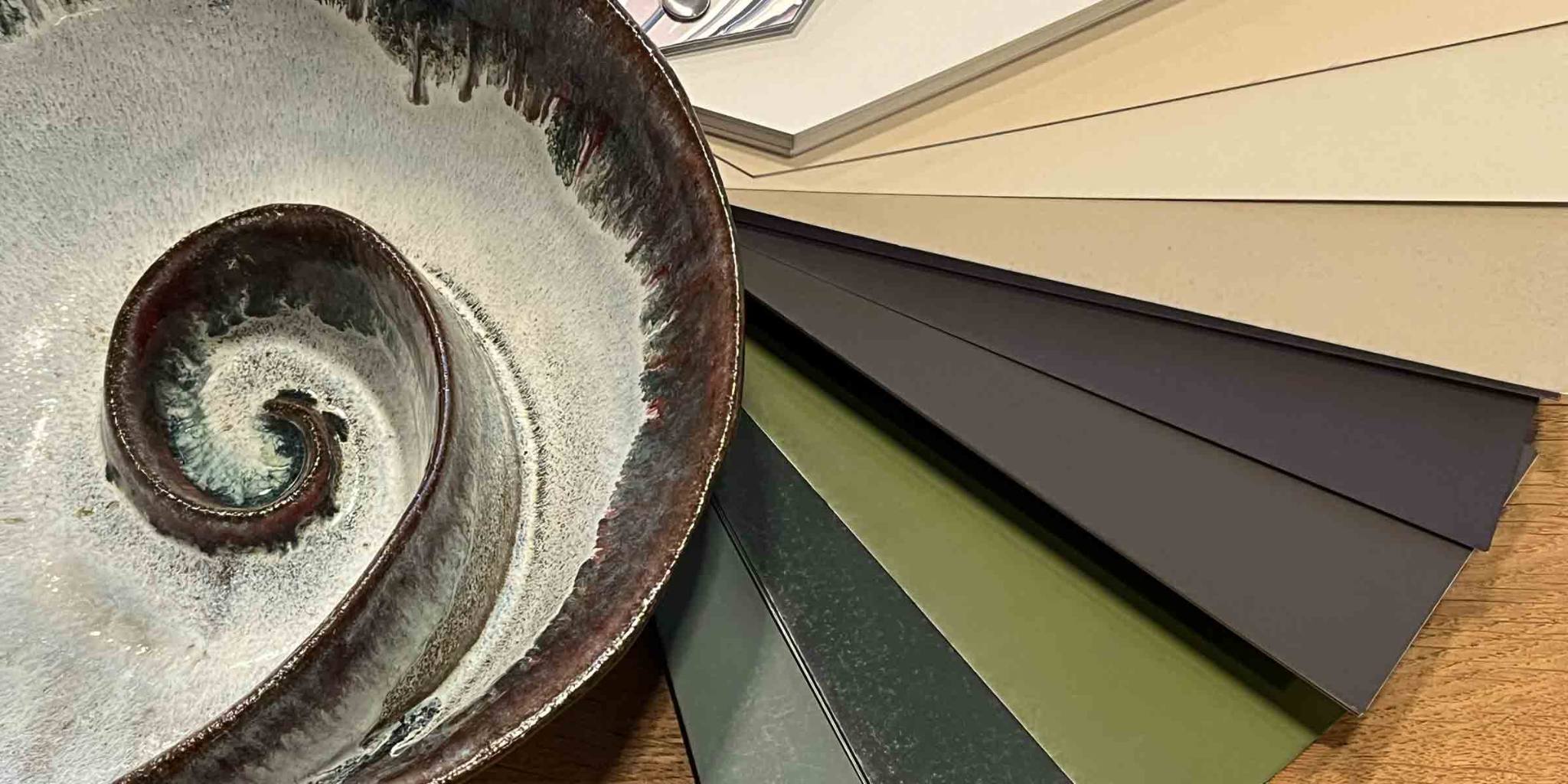 Close-up of a ceramic bowl with a spiral design in earthy tones, placed beside a selection of neutral and green-toned color swatches on a wooden surface.