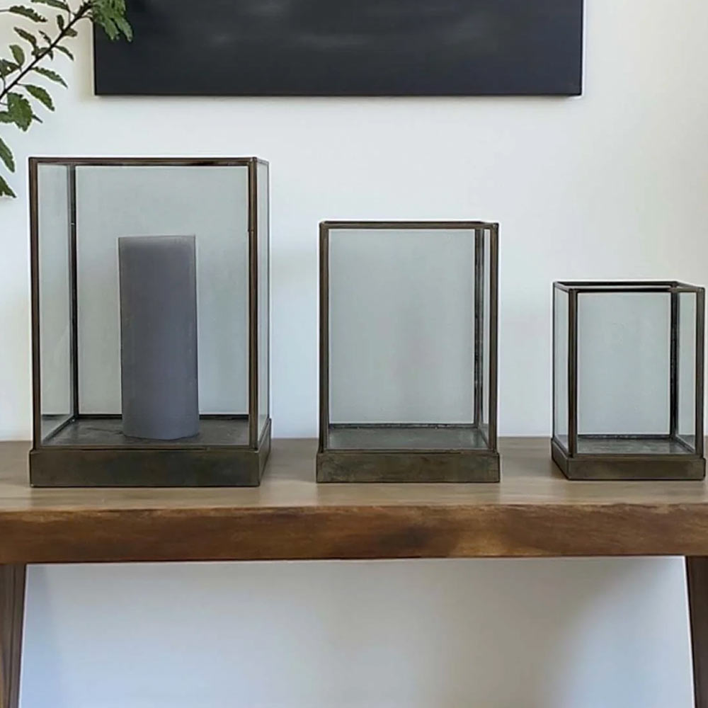 Three Aki Aged Hurricane Lanterns made from aged metal and clear glass on a wooden surface with a white wall background