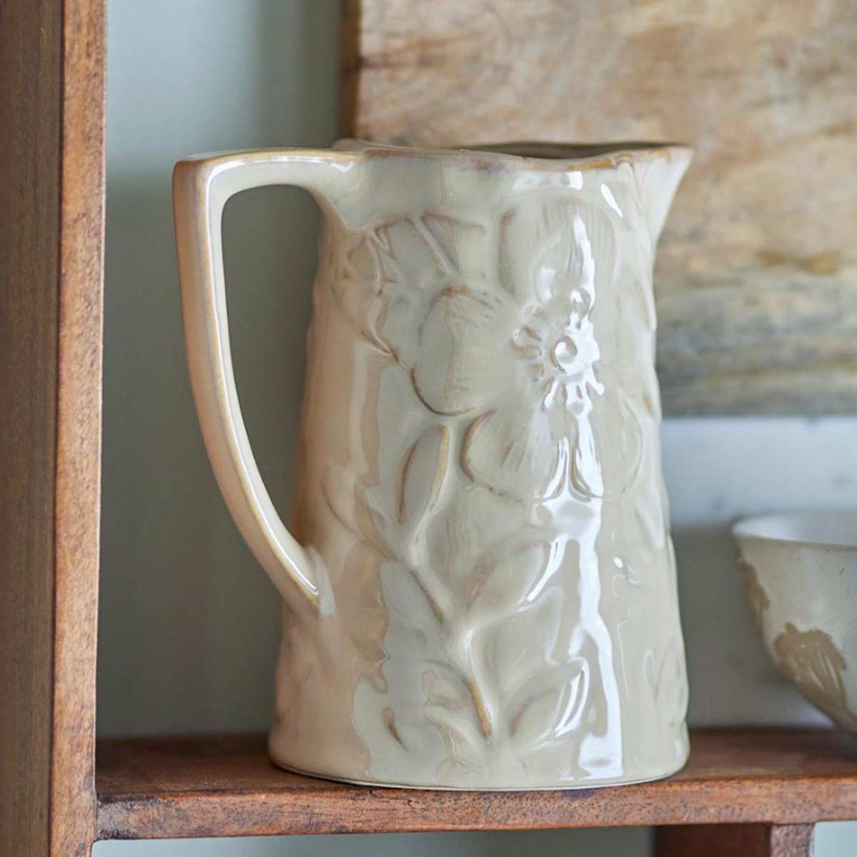 Aldon caramel-glazed ceramic jug with an embossed floral pattern, designed for serving drinks or displaying flowers - displayed on rustic wooden shelf with a stone wall behind.