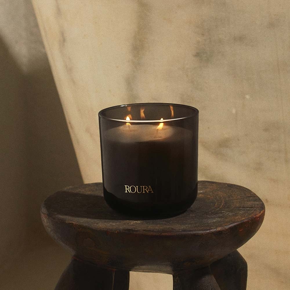 Roura Ancient Woods 3-wick candle in dark glass jar with deep woody scent on a wooden stool