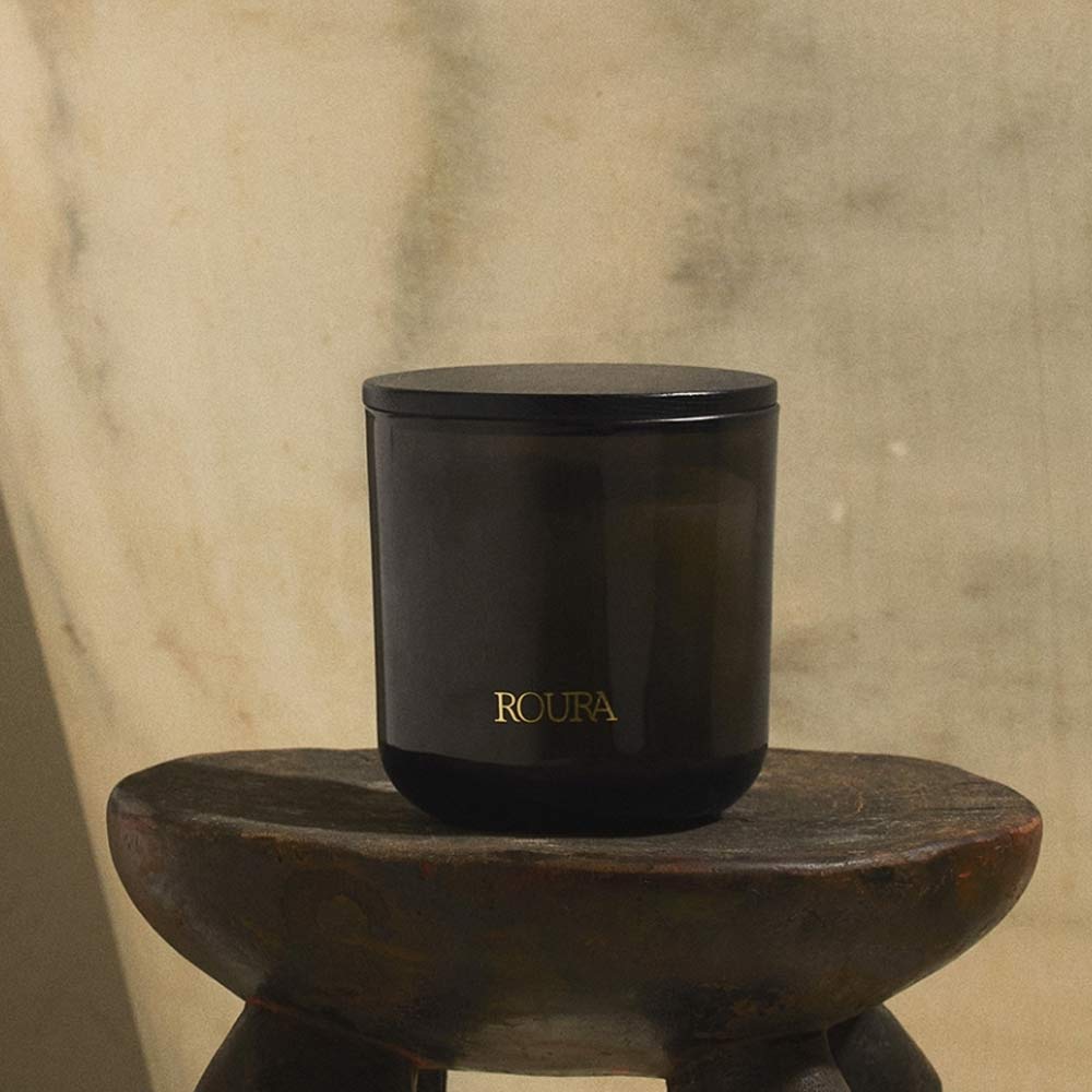 Ancient Woods Black Glass Jar Candle with lid on by Roura branding on a wooden stool against a textured beige wall.