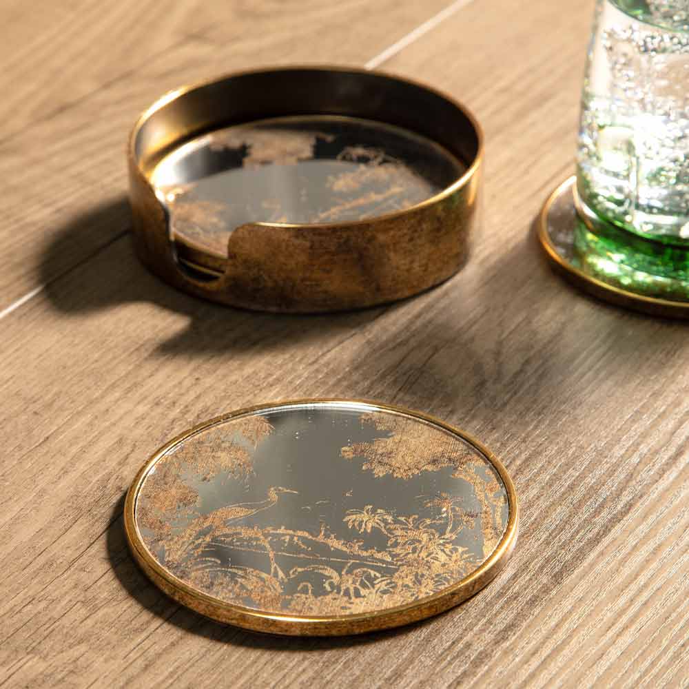 A round vintage-style brass and glass coaster with an etched landscape scene, shown with its matching circular holder on a wooden tabletop next to a green glass tumbler.