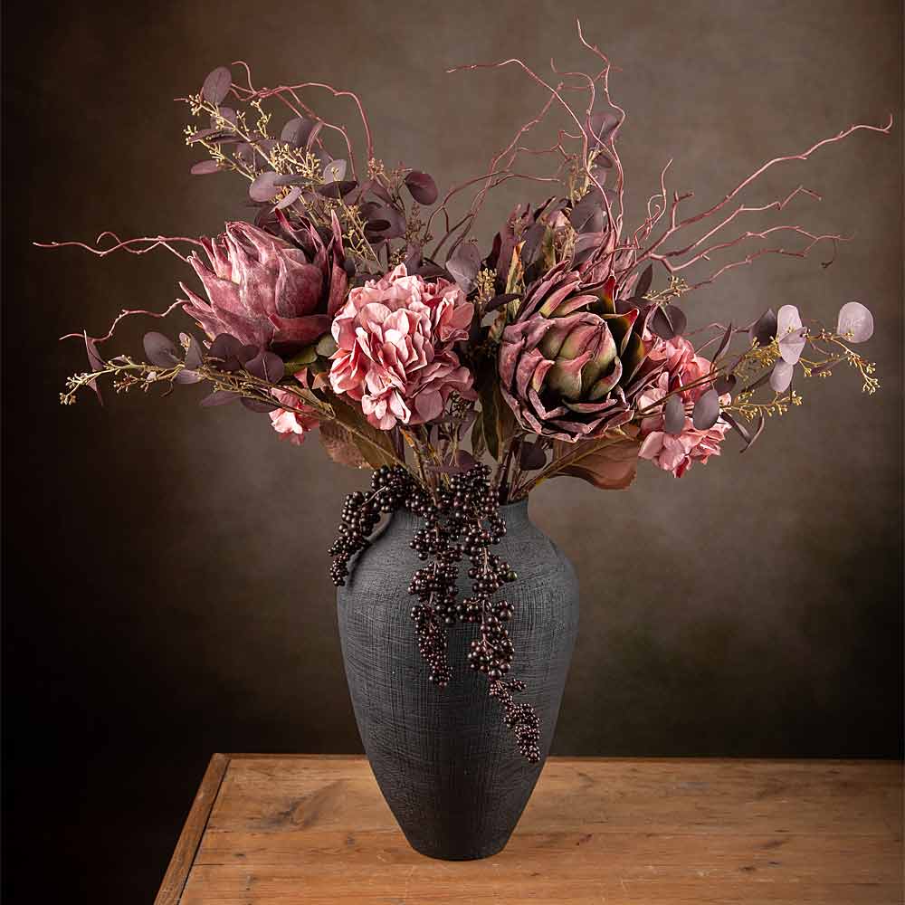 Faux Floral display containing rose pink hydrangea,Plum Protea, dogwood, aubergine eucalyptus and cotoneaster in a black textured vase on old wood table against a brown wall.