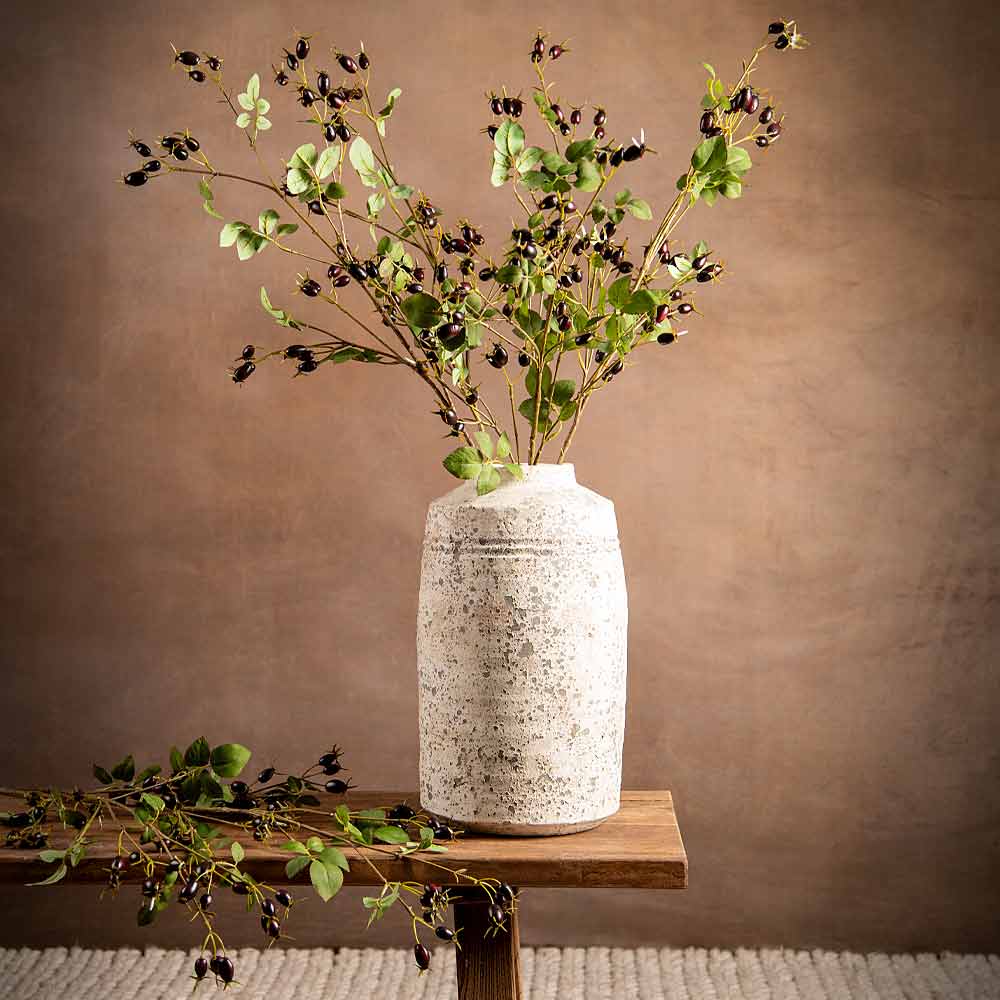 A speckled off-white ceramic vase filled with dried green faux foliage and small dark purple rose hip berries, displayed on a wooden table against a warm brown textured backdrop.