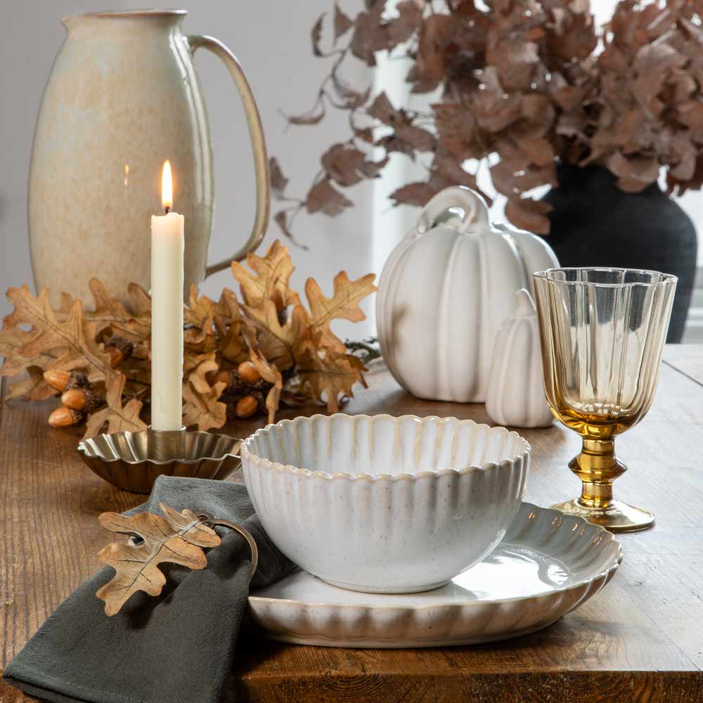A styled tabletop scene with a white scalloped ceramic bowl and matching plate set on a wooden table, surrounded by autumn decor including a candle, dried leaves, acorns, white pumpkins, and a ribbed clear glass goblet.