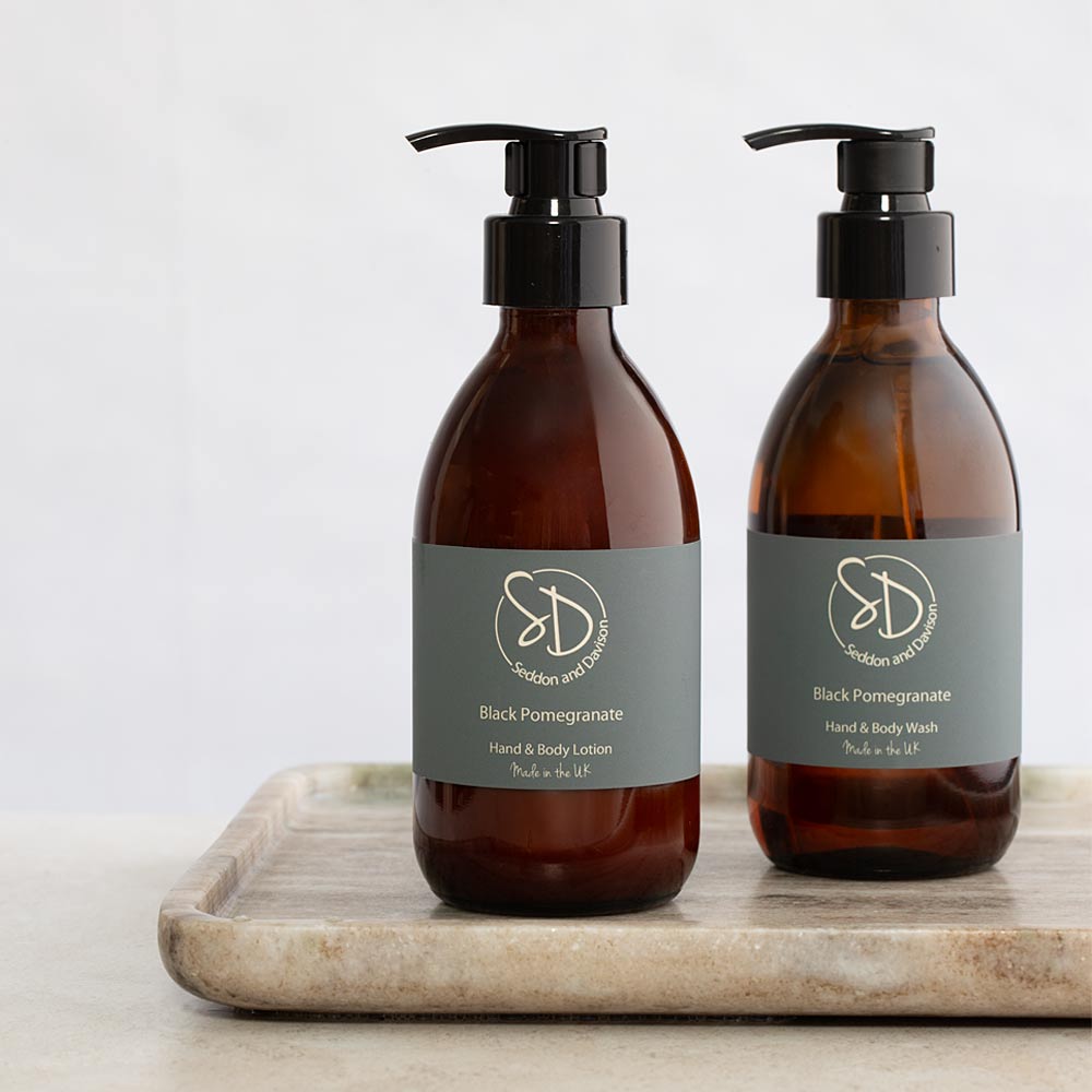 Two amber glass pump bottles with dark grey labels reading 'Black Pomegranate Hand & Body Lotion' and 'Black Pomegranate Hand & Body Wash' by Seddon and Davison, displayed on a marble tray against a light neutral background.