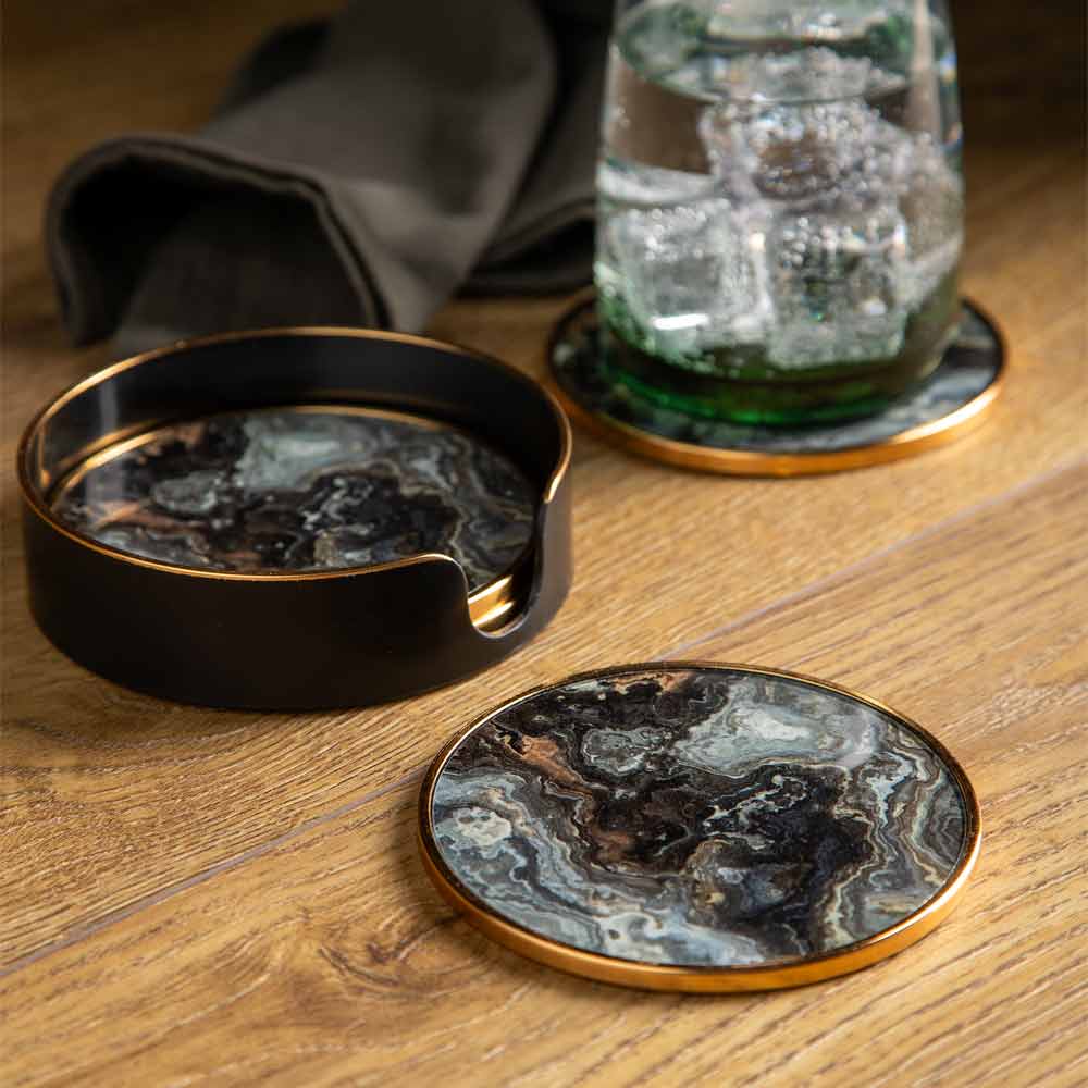 A set of round coasters with a black, brown and gray marbled pattern and gold trim, shown on a wooden table with a dark grey cloth and a glass of sparkliong water in the background.