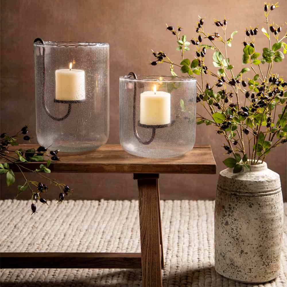 Two clear textured glass candle holders with black metal candle sleeves, each containing a lit cream pillar candle, displayed on a wooden bench in a styled interior setting with faux greenery and a distressed ceramic vase.