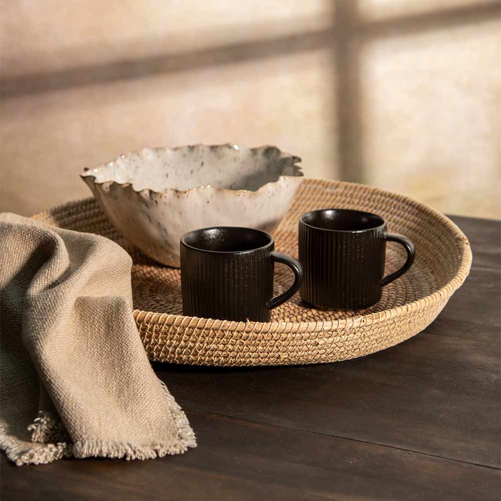 Two black ribbed ceramic mugs with handles sit on a round woven tray, alongside a speckled beige bowl and a draped beige cloth, set on a dark wooden table with warm natural light casting soft shadows.