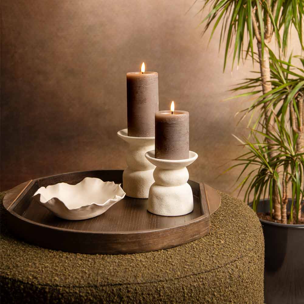 Two lit brown pillar candles on white ceramic candle holders on a round wooden tray, set on a textured olive-green ottoman with a potted plant in the background.