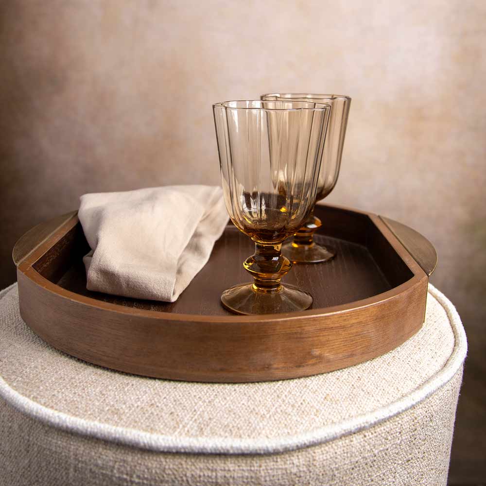 A round wooden serving tray with a folded cream linen napkin and two amber-tinted stemware glasses, displayed on a textured beige upholstered stool against a warm neutral wall.