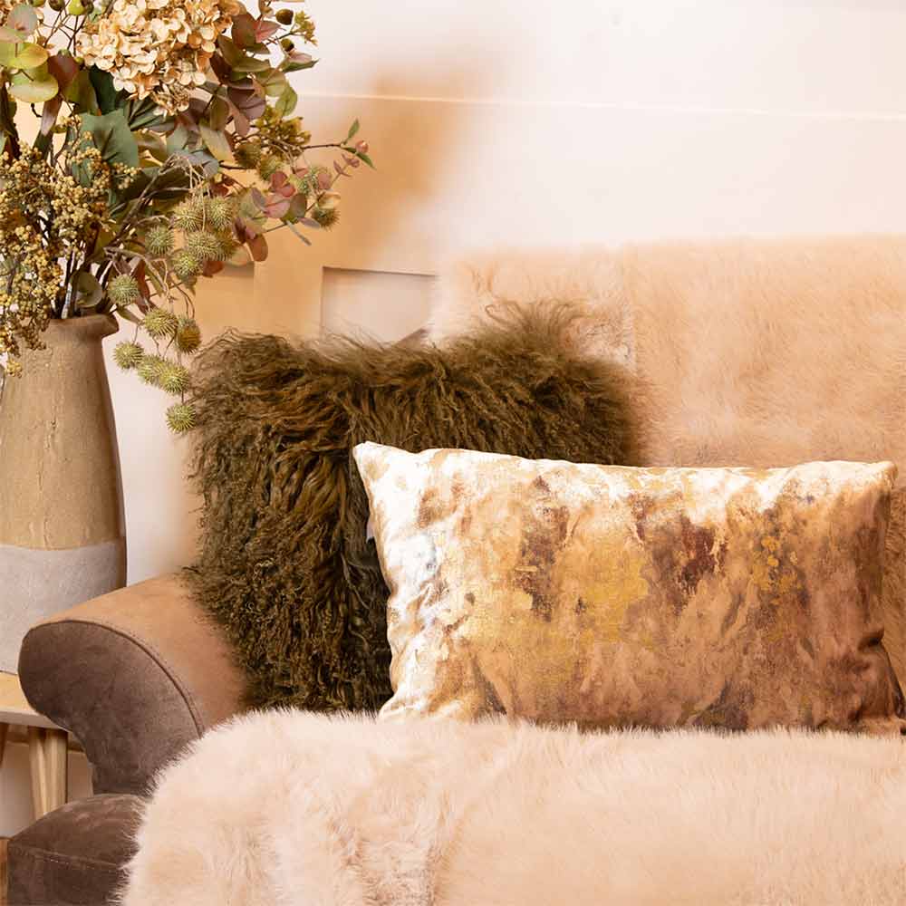 Oblong Caesium cushion with a gold abstract metallic front and olive green velvet reverse - styled with other cushions on a sofa with a vase of flowers in the background