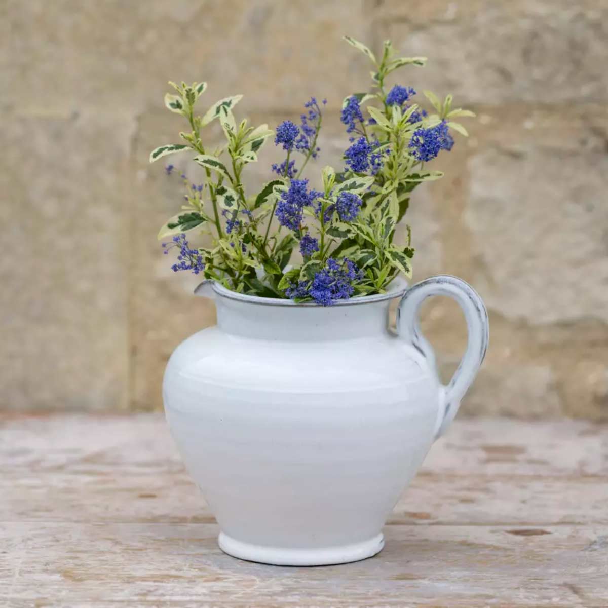 Carla White Glazed Jug – hand-thrown ceramic jug with food-safe white glaze and wildflowers, styled on a weathered wooden table against a stone wall