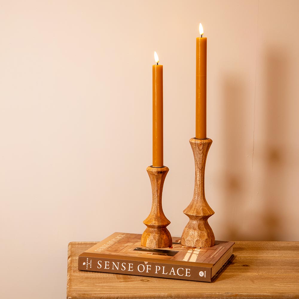 Two lit candles in Large and Small Cirilo Natural Wood Candlesticks placed on a book on a rustic wooden surface