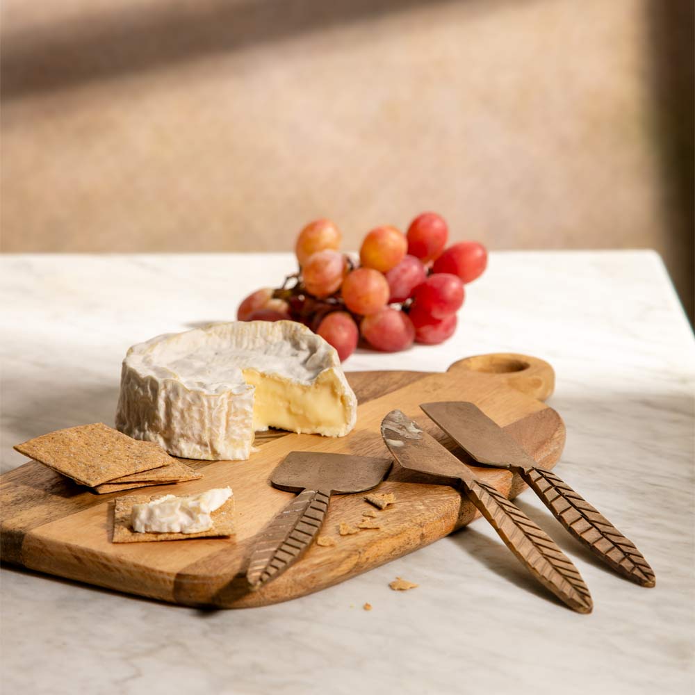 A wooden cheese board with carved round handle, styled with a wheel of brie, cheese knives, crackers, and a bunch of red grapes on a light marble surface.