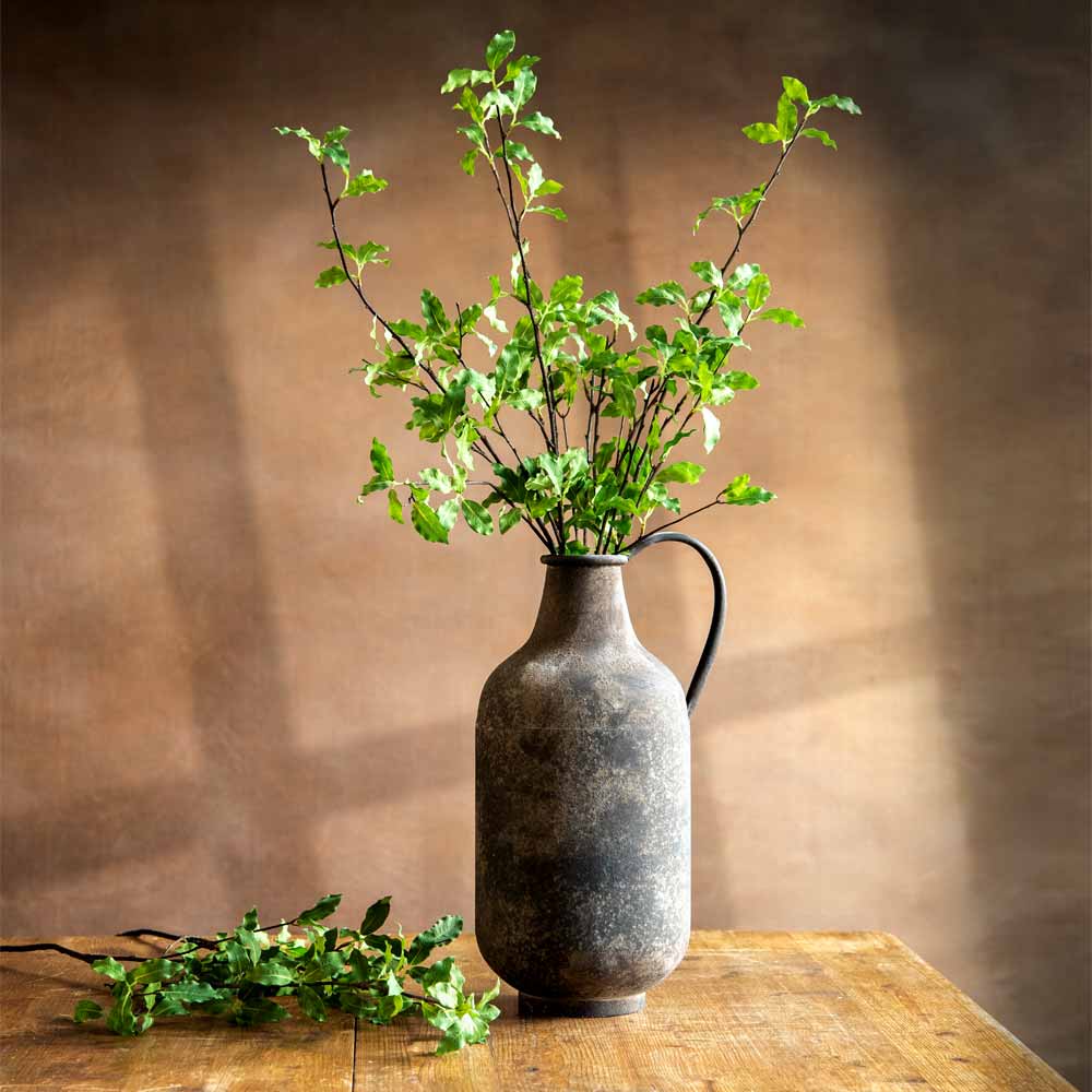 A rustic dark gray ceramic pitcher vase with a handle, filled with green leafy branches, set on a wooden table against a warm brown textured background.