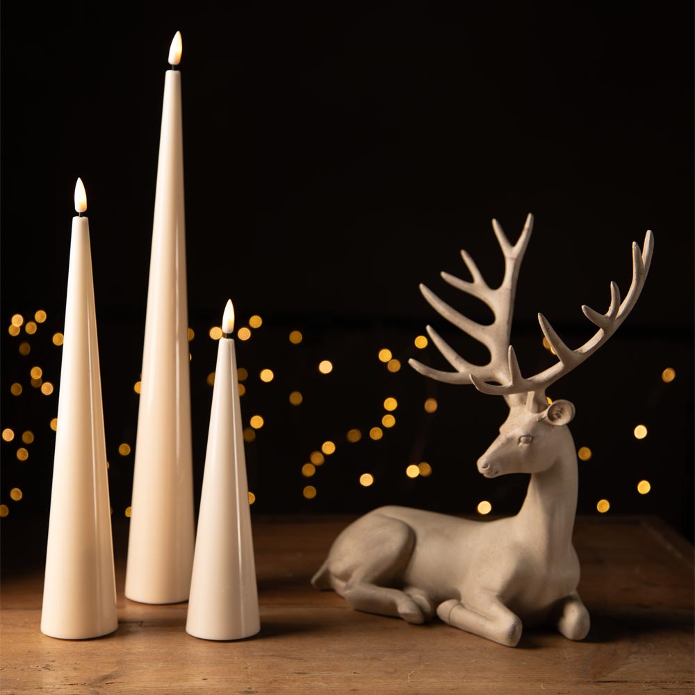 Three LED tapered cream pillar candles with flame tips stand on a wooden surface beside a cream-coloured deer figurine with large antlers, set against a dark background with warm blurred bokeh lights.