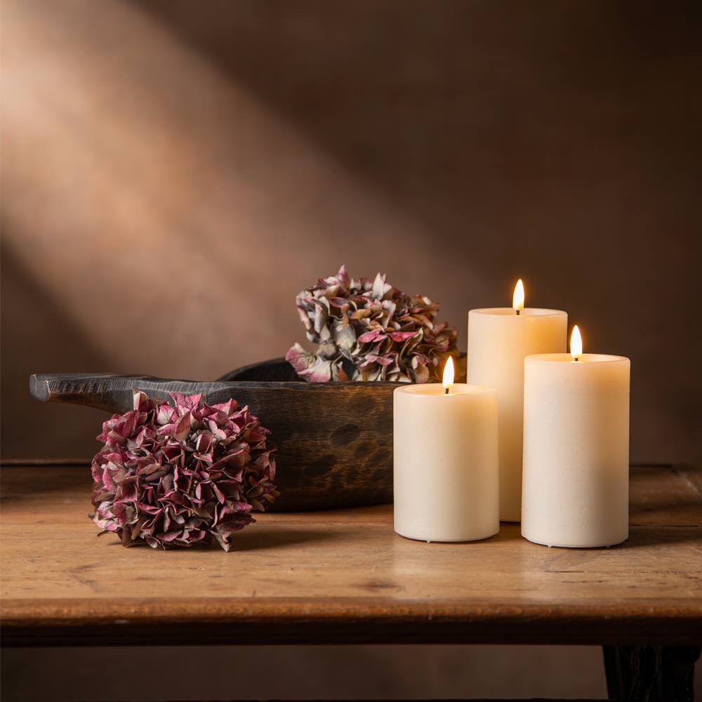 Three lit cream LED pillar candles on a rustic wooden table, styled with dried pinkish-purple flowers and a dark wooden bowl against a warm brown background.