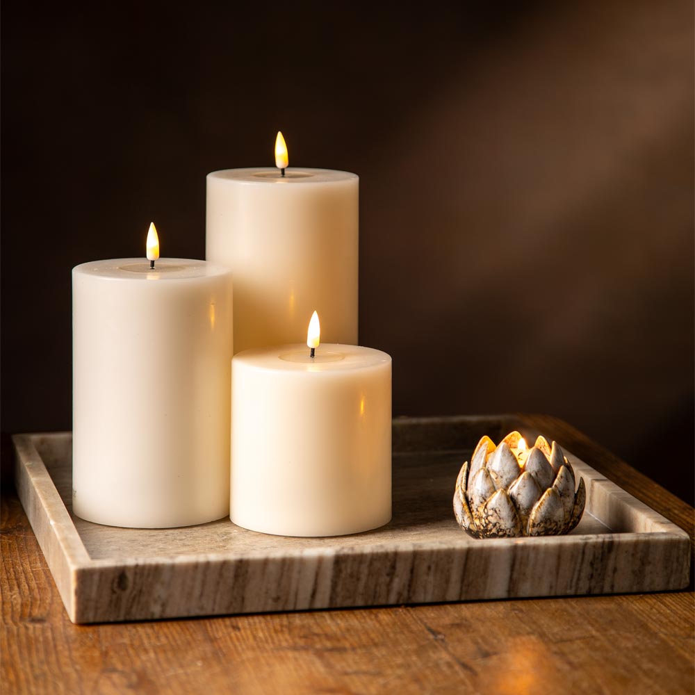 Three lit cream pillar candles arranged on a marble tray next to a small lotus-shaped candle holder on a wooden table against a dark brown background.