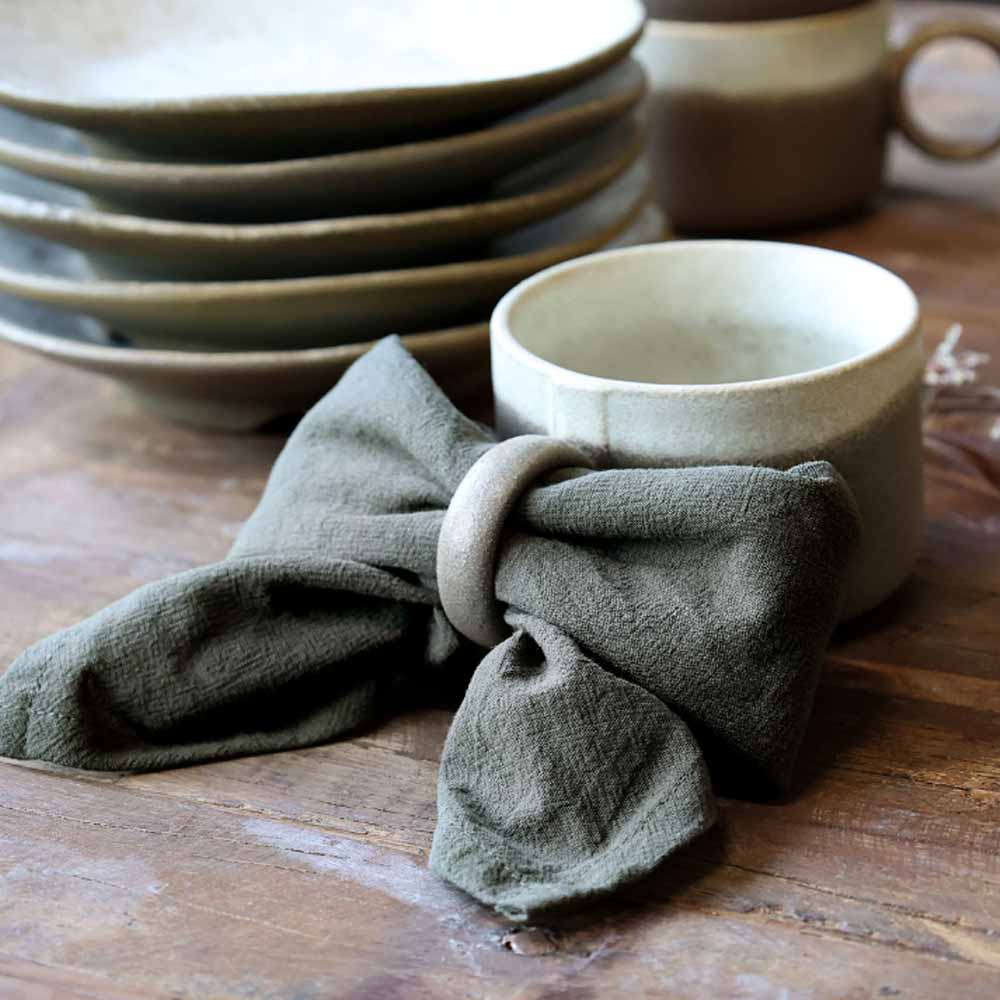 Valence Ceramic Mug with a Dark Olive Cloth Napkin on a wooden surface, with stacked ceramic plates in the background.