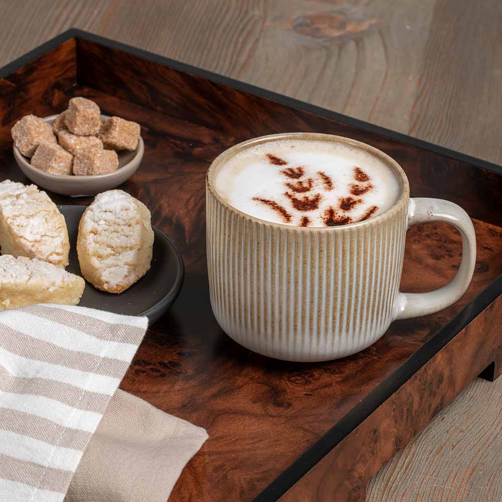 A ribbed ceramic mug filled with a frothy latte sits on a dark wooden tray, alongside small pastries, sugar cubes, and a striped napkin on a wood surface.