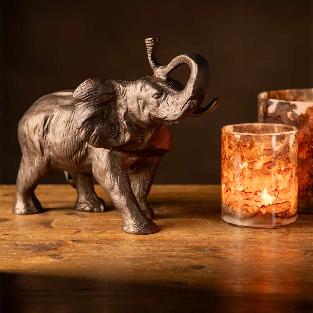 A metallic elephant figurine with a raised trunk stands on a wooden surface next to a glowing glass candle holder against a dark brown background.