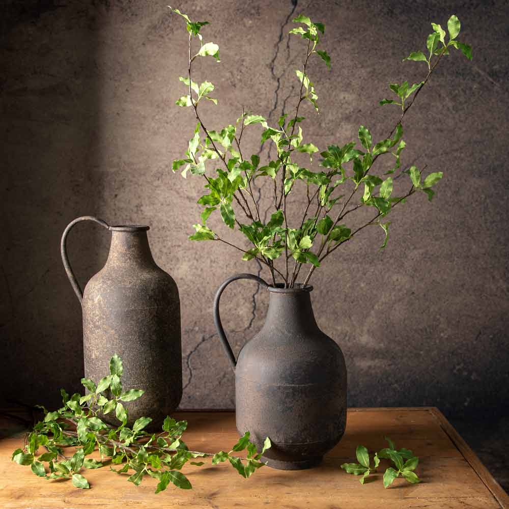 Two dark rustic metal pitchers with handles sit on a wooden table against a distressed beige wall. One pitcher is empty, and the other is filled with leafy green branches.