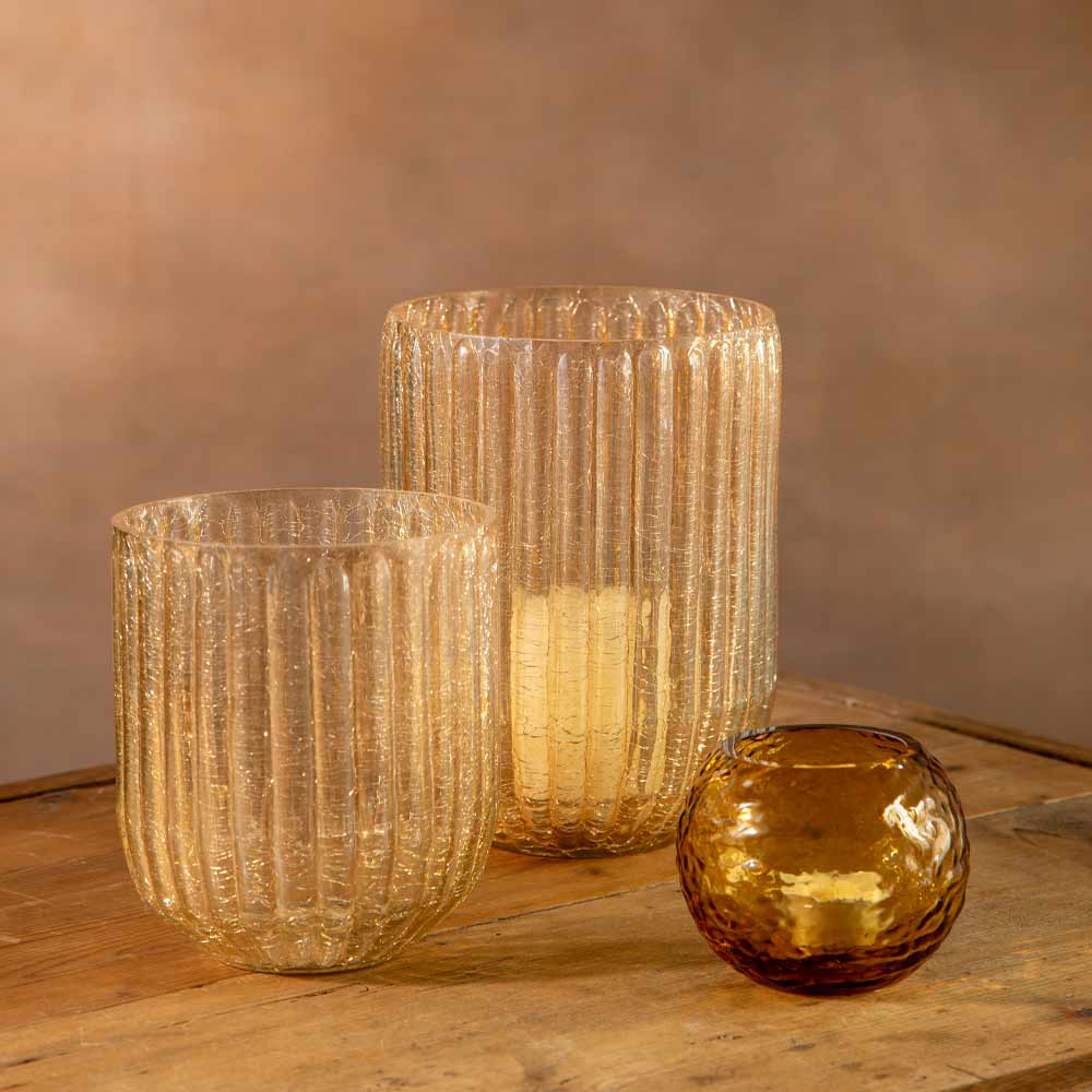 Two ribbed glass candle holders in clear crackle and amber tones arranged alongside an amber glass tealight holder on a wooden surface against a warm brown background.