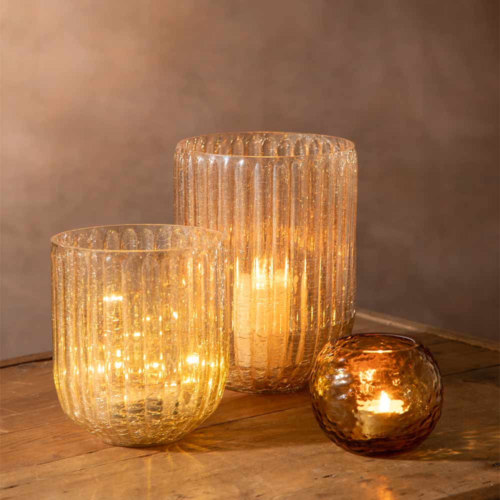 Two amber glass candle holders with ribbed and hammered textures, lit one with a warm pillar candle and another with a led light string. alongside a small amber l tea light candle glows softly these are arranged on a wooden surface against a neutral brown background.