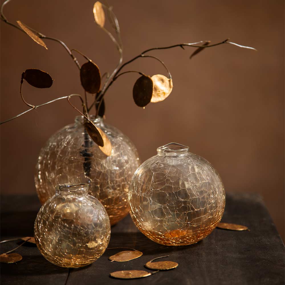 Three round crackle-glass vases in clear and warm amber tones, with one larger vase holding a decorative branch of metallic gold seed pods, set on a dark surface against a warm brown background.