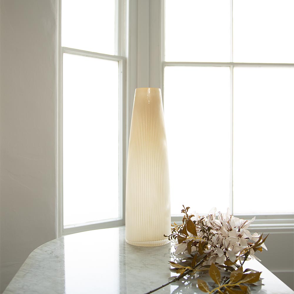 A tall, ribbed cream-coloured ceramic vase stands on a light marble table in front of bright white window panes, with faux flowers placed beside it.