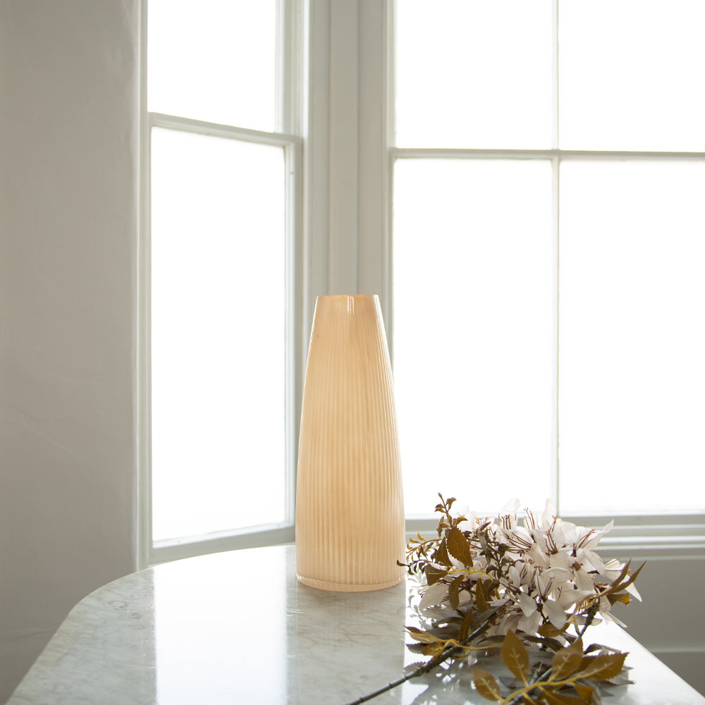 A ribbed, tapered ceramic vase in a warm cream-beige colour stands on a light marble table in front of a bright window, with white flowers and green leaves placed beside it.