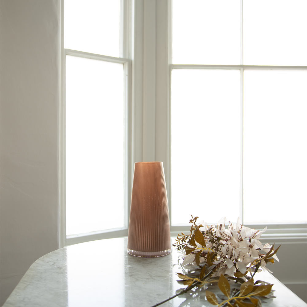 A ribbed, tapered ceramic vase in a warm dusty rose colour, standing on a white marble table next to dried white flowers in front of a bright, sunlit window.