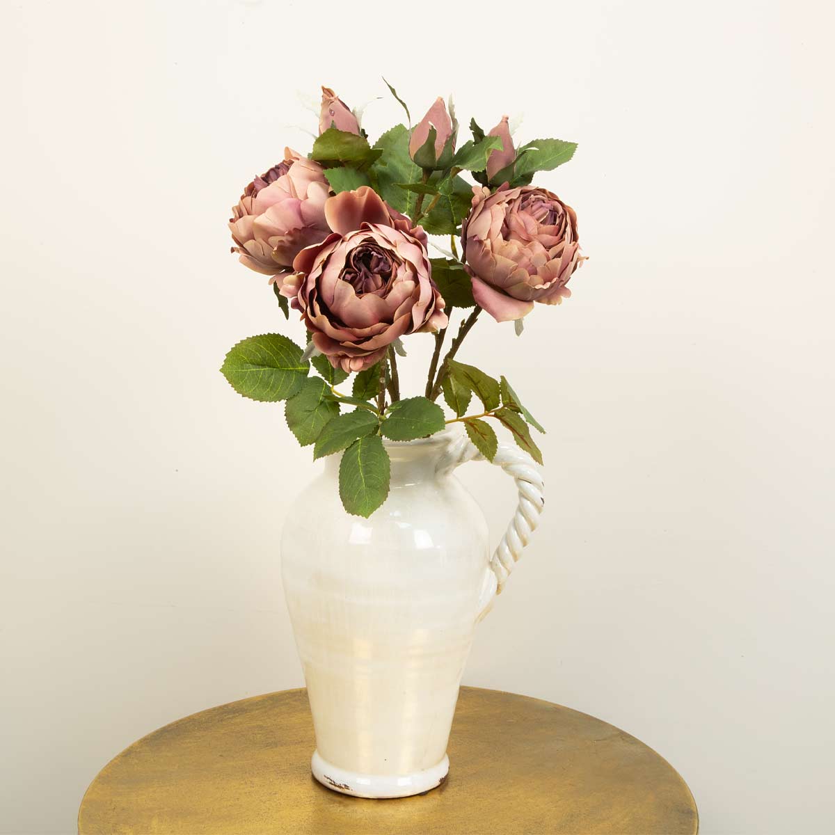 A bunch of artificial dark pink peony spray flowers arranged in a white vase, placed on a gold surface against a white background.