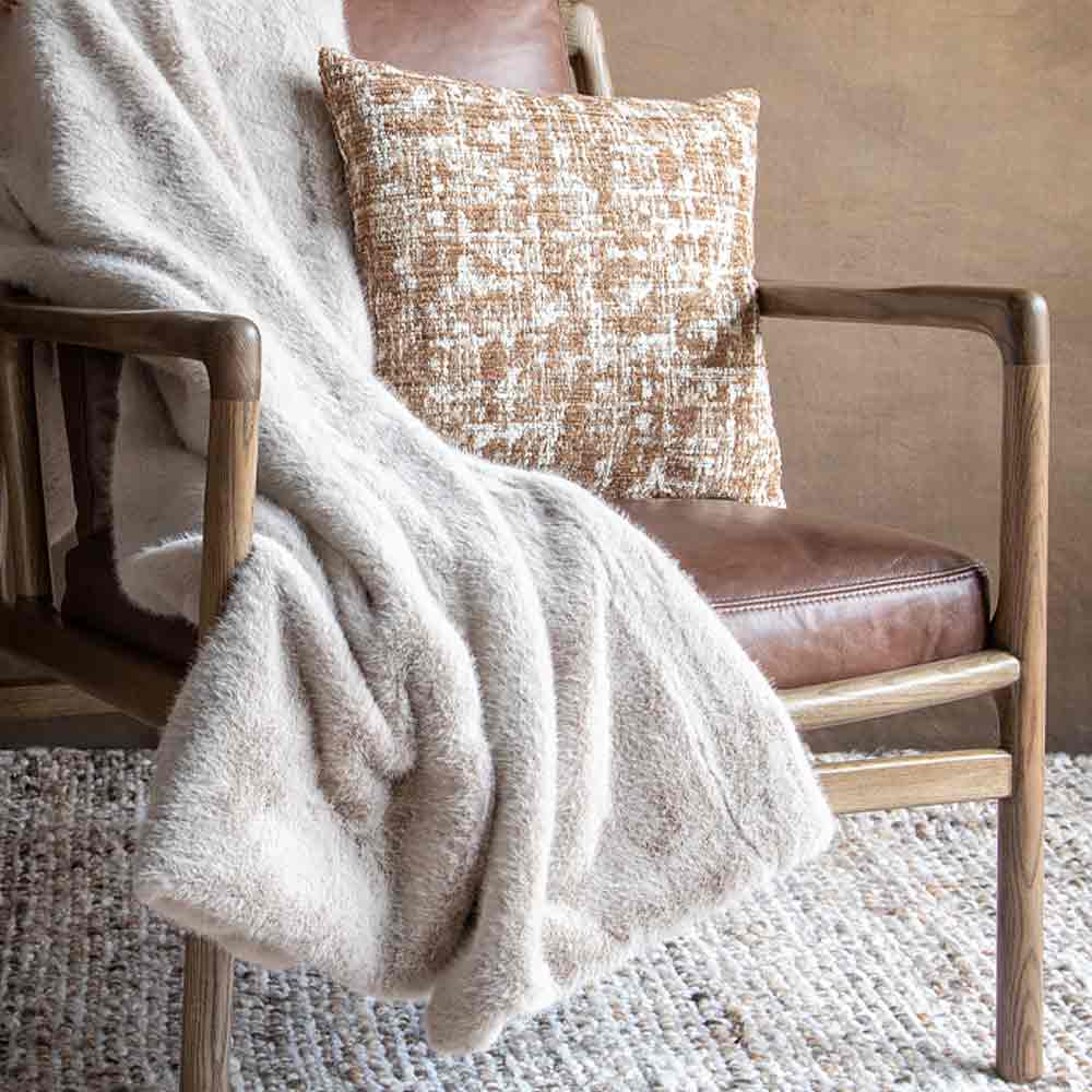A cozy living room scene featuring a plush, textured cream throw blanket draped over a mid-century modern wooden armchair with a brown leather seat. with a textured cream and brown pillow rests on the chair.
