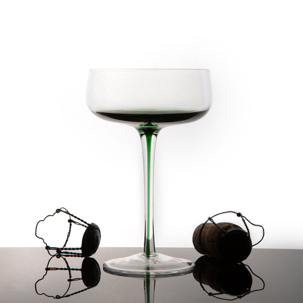 A coupe-style glass with a green-tinted lower portion of the bowl thar proceeds into the stem , shown on a reflective black surface with two dark bottle stoppers nearby against a white studio background.