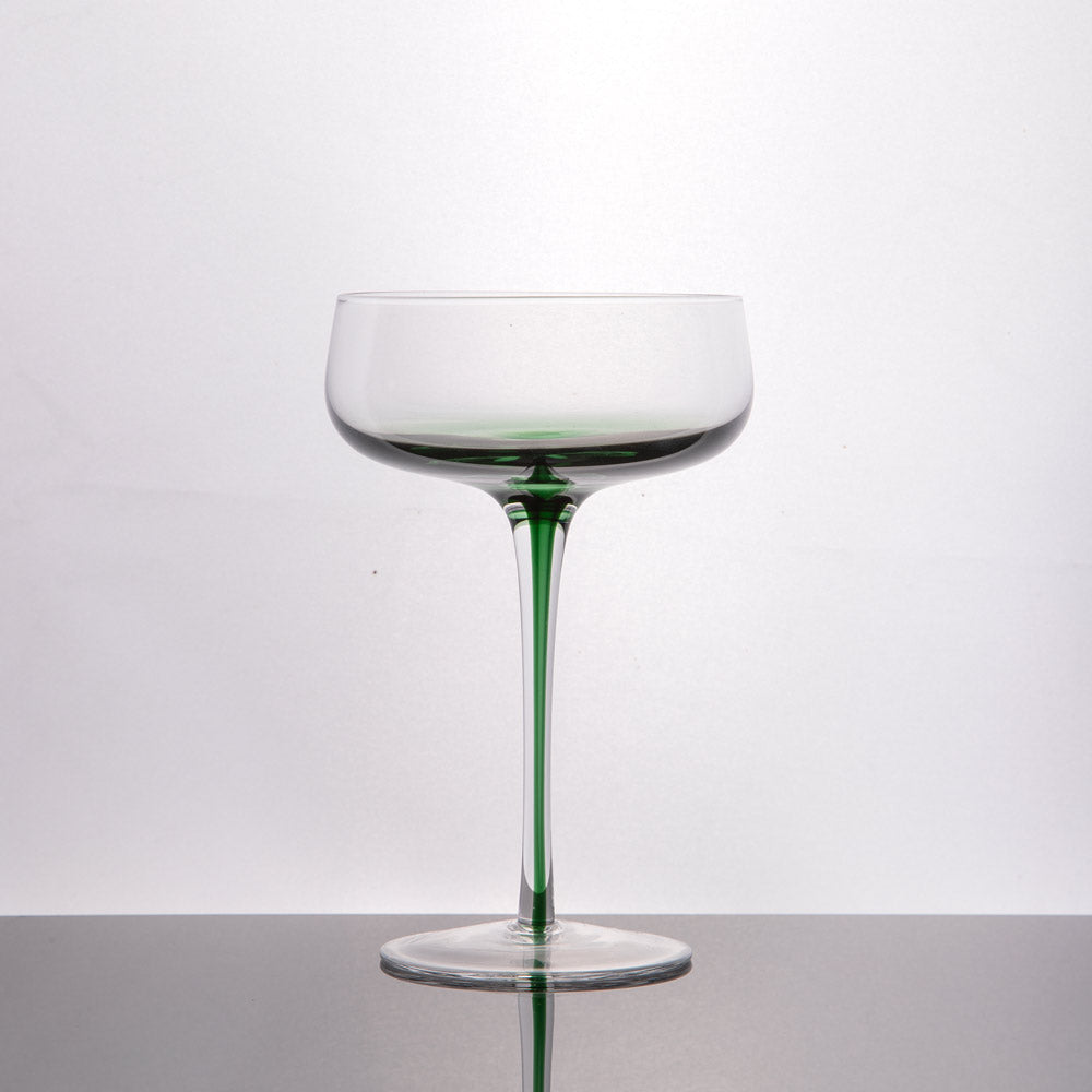 A clear stemmed glass with a green-tinted base and stem, photographed on a reflective dark surface against a light gray studio background.