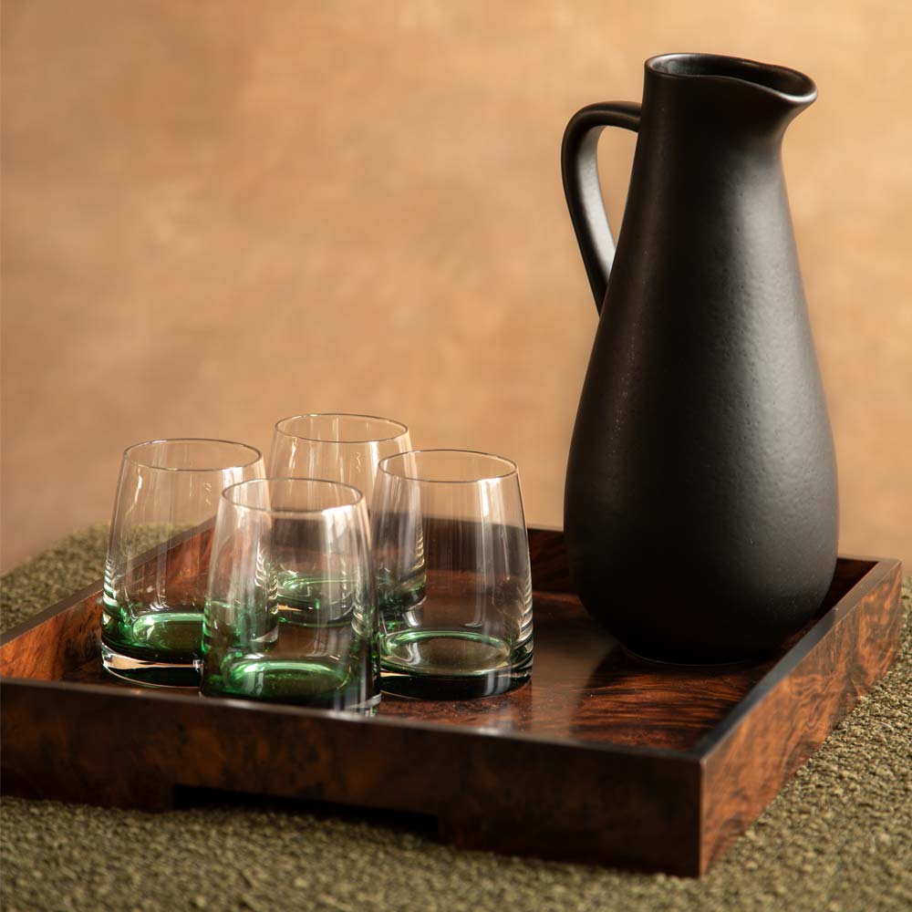 A matte black ceramic carafe and four tumbler glasses with a translucent green base arranged on a dark wooden serving tray against a warm beige background.