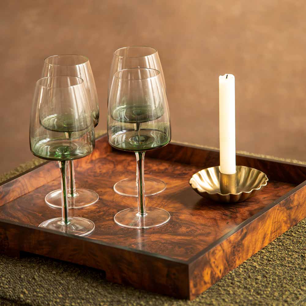 Four stemmed wine glasses with a green tinted glass base, arranged on a Square wooden serving tray alongside a white dinner candle in a scalloped brass holder, set against a warm brown background.