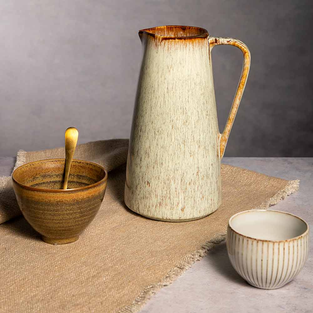A rustic ceramic pitcher with a cream and brown speckled glaze stands on a burlap cloth, alongside a small ribbed bowl with a spoon and a matching cup, against a neutral gray background.
