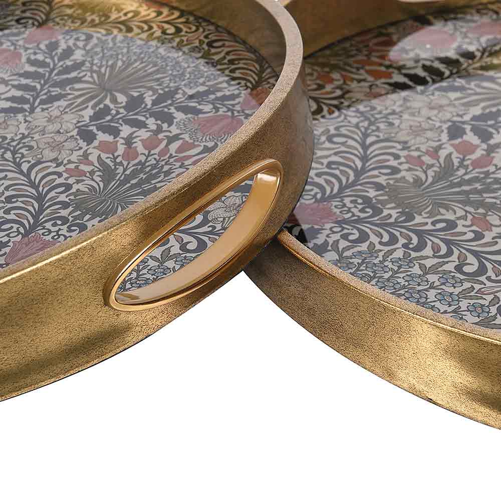 Close-up of round decorative trays with metallic gold-toned rims and handles, featuring a floral and botanical patterned surface in muted blue, gray, pink, and beige tones.