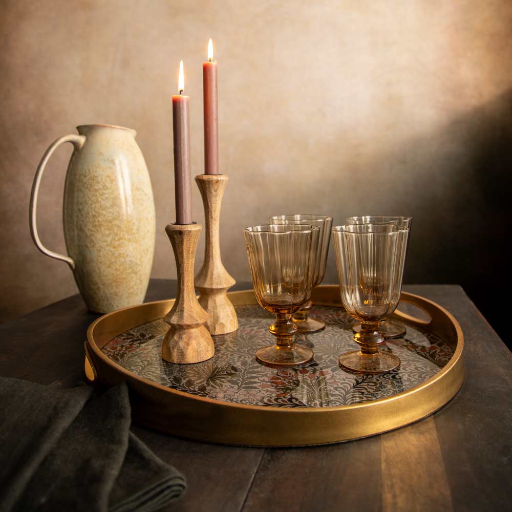 A styled tabletop scene with a round matt gold tray holding four goblet style glasses, two wooden candlesticks with lit dinner candles. A beige ceramic pitcher sits behind on a dark wood surface against a warm textured wall.