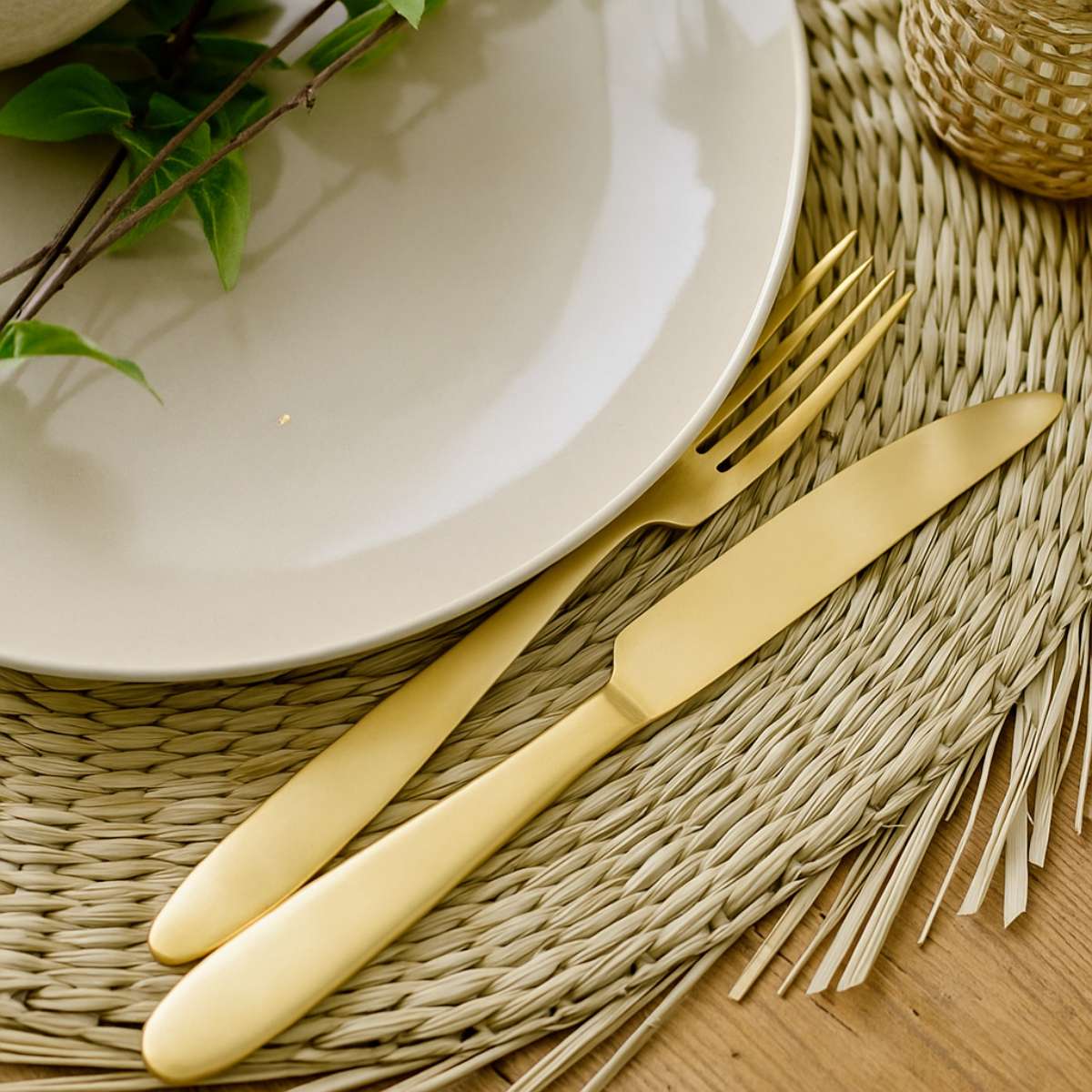 Frea Gold knife and fork set on a woven placemat with a white plate and greenery.