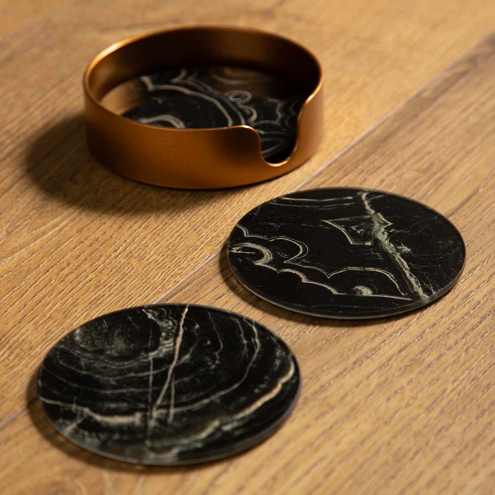Two round coasters on a light wood tabletop, including two black stone coasters with white marbled veins and one metallic bronze ring-shaped coaster holder.