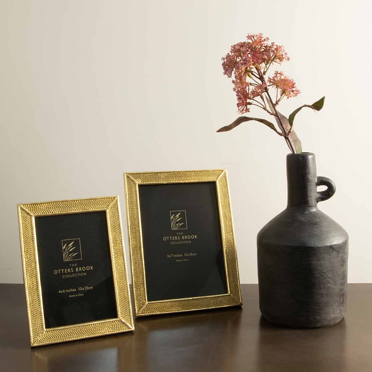 Gold dotted effect photo frames in 18x13cm and 20x16cm sizes styled on a dark wooden surface with a black vase and pink floral stem.