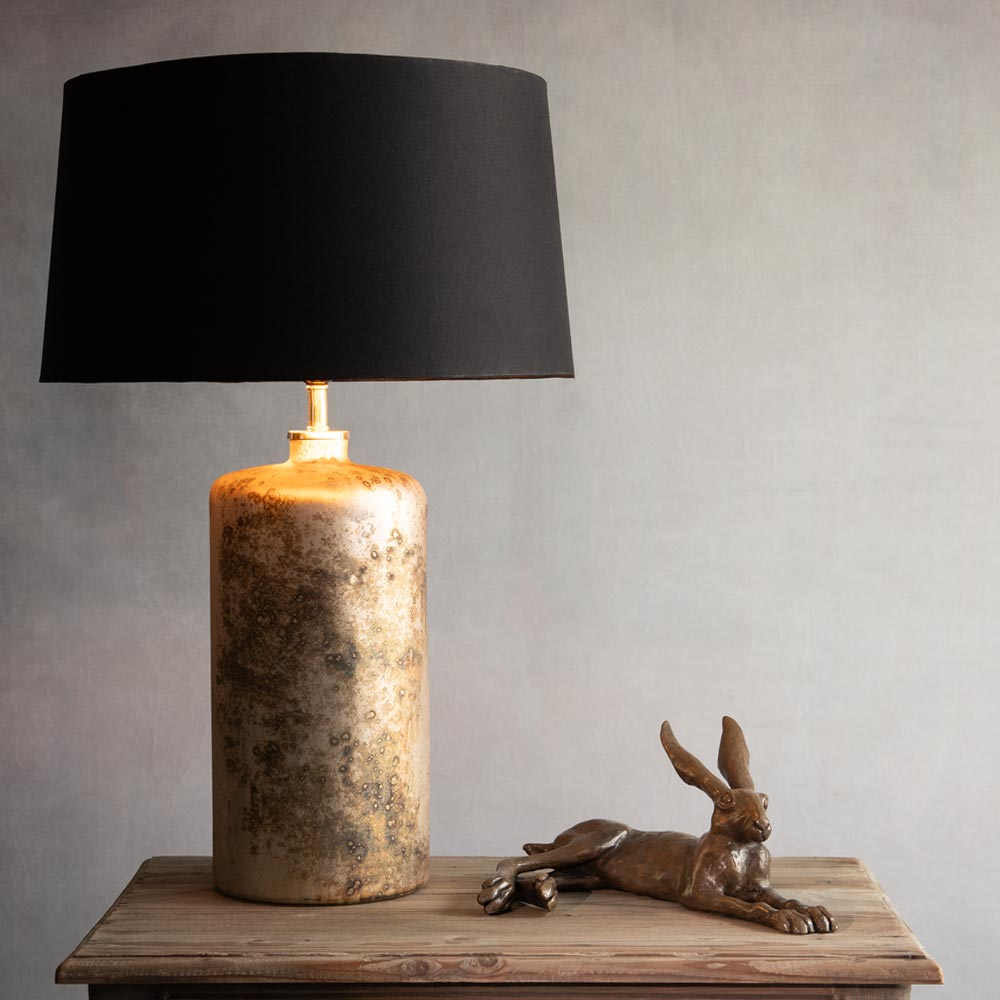 A table lamp with a black drum shade and a textured metallic gold base, styled on a rustic wooden table next to a bronze hare figurine against a neutral gray background.