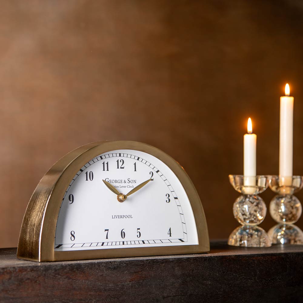 A George & Son Liverpool desk clock with a brass semi-circular frame and white dial, styled on a dark wooden surface next to two lit candles in glass holders against a warm brown background.