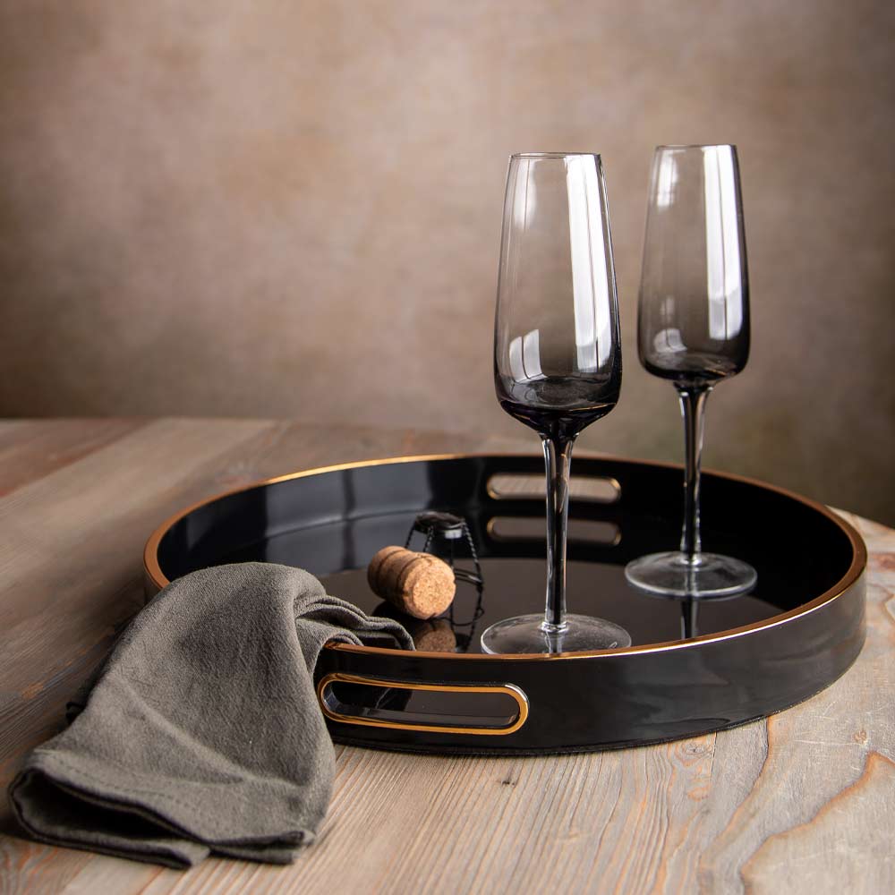 A round black serving tray with gold trim and cutout handles sits on a wooden table, styled with two dark tinted champagne flutes, a folded gray cloth, and a cork.