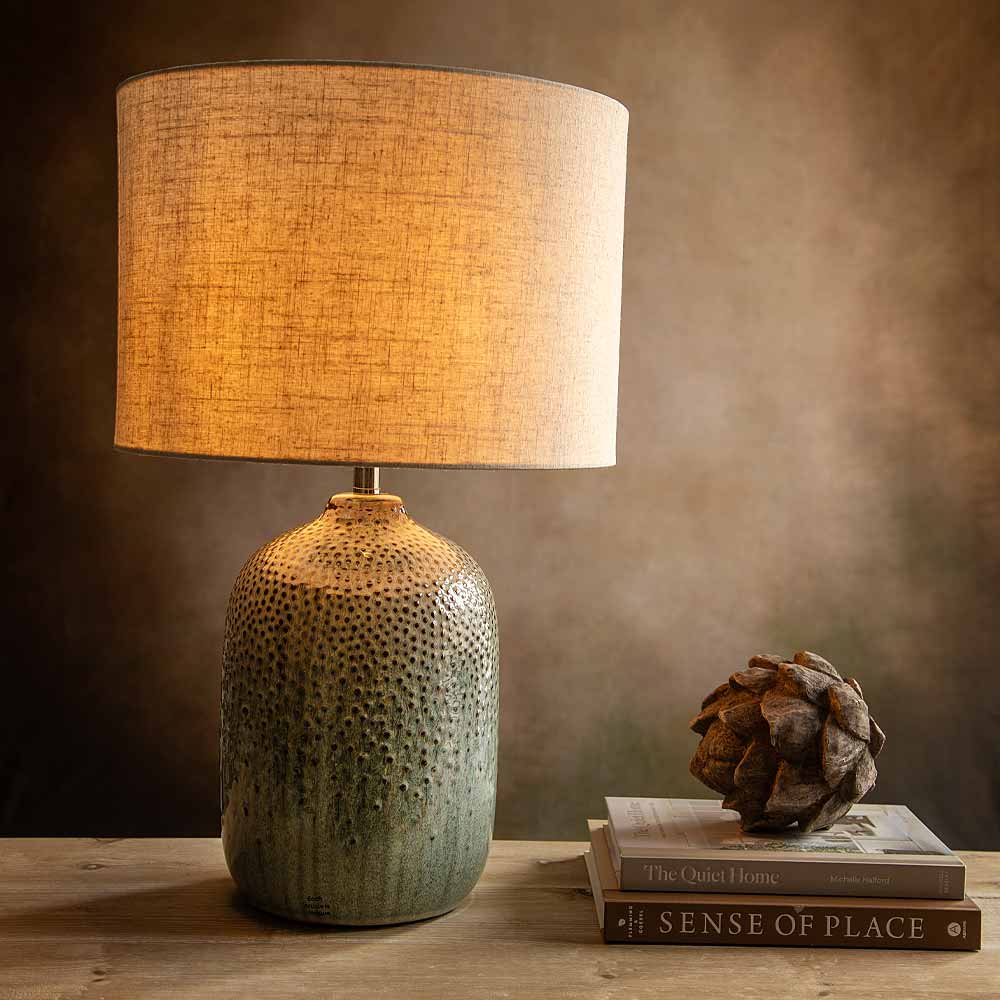 A table lamp with a textured, dimpled ceramic base in a muted green-gray and a warm beige linen drum shade, lit to cast a warm glow. The lamp sits on a rustic wooden table next to a stack of books and a decorative pinecone, set against a dark, warm brown background.