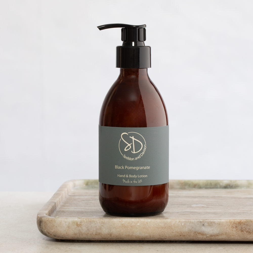 An amber glass pump bottle of Seddon and Davison Black Pomegranate Hand & Body Lotion, resting on a light marble tray against a soft white background.